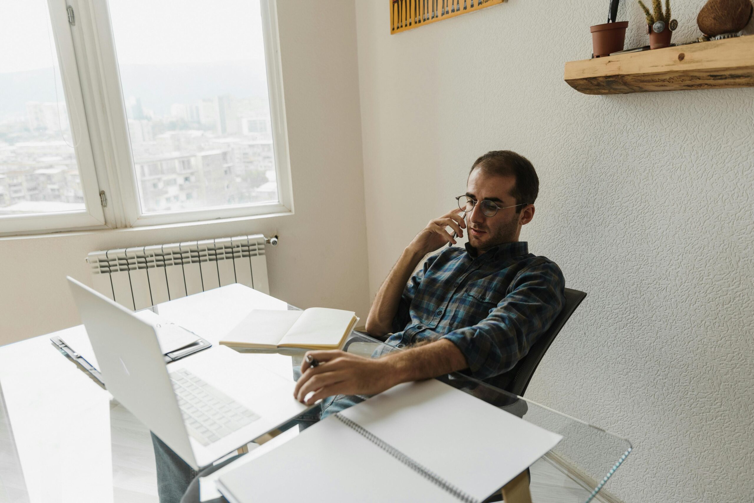 A young man working remotely in his home office, talking on the phone, using a laptop.